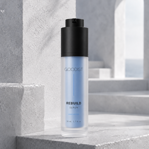 A bottle of GOODIST REBUILD Serum with a black pump and a frosted light blue bottle, set on a gray concrete staircase in a minimalist architectural setting.