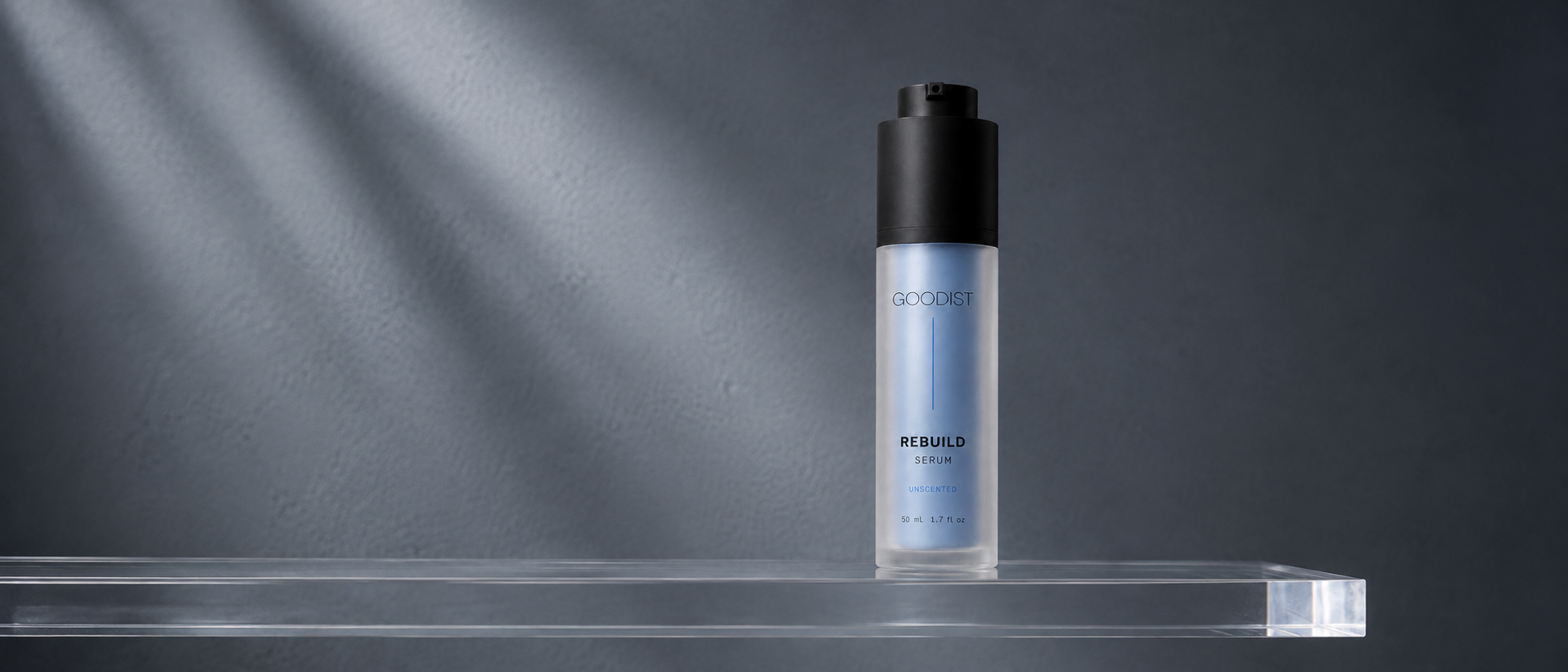 A bottle of GOODIST REBUILD face serum stands on a clear acrylic platform against a dark gray background with dramatic diagonal light rays.