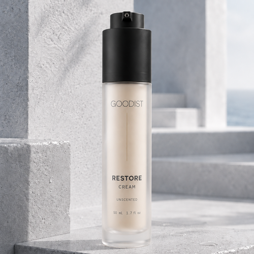A 50 mL bottle of GOODIST Restore Cream, unscented, standing on a gray stone ledge with a blurred architectural background.