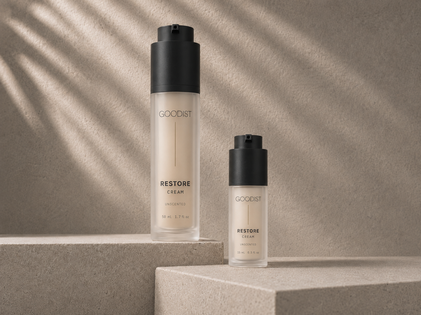 Two GOODIST Restore Cream bottles, one larger pump bottle and one smaller airless pump bottle, displayed on a minimalist concrete pedestal with soft leaf shadows.