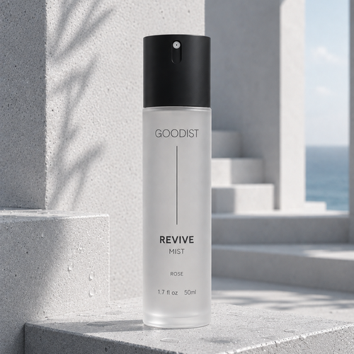 A 50ml bottle of GOODIST Revive Mist in Rose, standing on concrete steps with a blurred outdoor background showing a sea view and shadows.