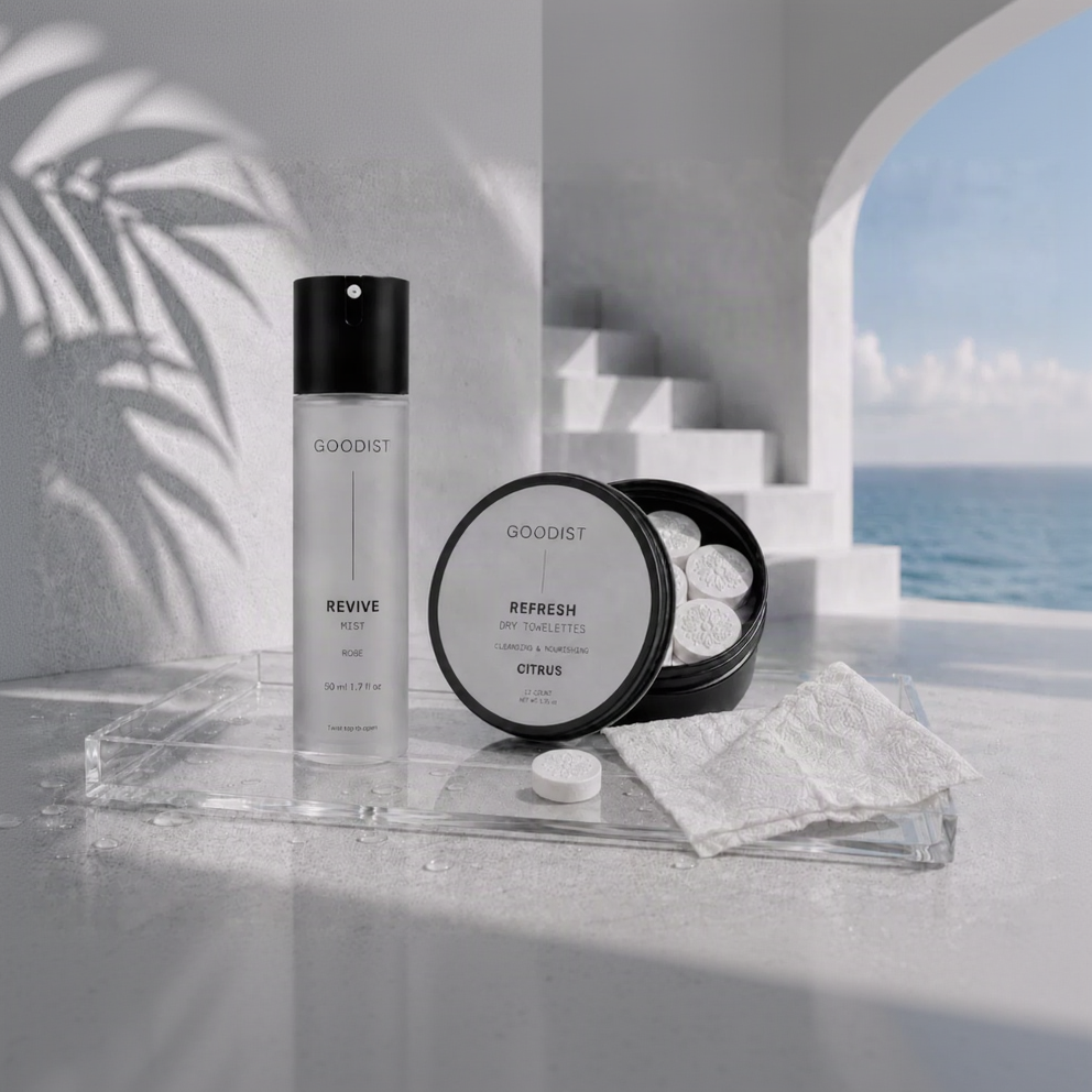 Goodist skincare set featuring a Revive Mist bottle and a Refresh Dry Towelette pack, styled on a clear acrylic tray with a white towel and a single towelette disc, set against a modern white architectural background with a glimpse of the ocean.